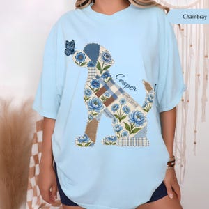 May include: A light blue t-shirt featuring a graphic of a dog composed of patchwork patterns, including blue floral prints, brown solid sections, and blue plaid squares. A small blue butterfly sits on the dog's nose, and the name Cooper is printed in blue script.