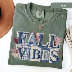 May include: A moss green t-shirt featuring the text "FALL VIBES" in a patchwork design. The letters are blue and beige, with plaid and leaf accents. A gold necklace with a pendant is draped on the shirt.