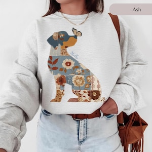 Puede incluir: Una sudadera de cuello redondo color gris ceniza con el gráfico de un perro sentado. El cuerpo del perro está compuesto por varios patrones florales de estilo patchwork en tonos azul, marrón y crema, con una pequeña mariposa en la cabeza.