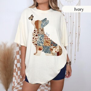 May include: An ivory short-sleeve t-shirt featuring a large graphic of a dog silhouette filled with a patchwork pattern of blue, brown, and white floral and leaf prints. A small butterfly sits on the dog's nose, and the name Molly is printed in script.