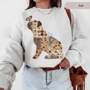 Op de afbeelding: Een asgrijs sweatshirt met lange mouwen en een patchwork-hondensilhouet gevuld met herfstachtige bloemen- en bladpatronen in bruin-, oranje- en crèmetinten, met een vlinder op de kop en de naam Molly in het ontwerp verwerkt.