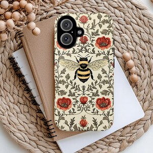 May include: A phone case with a floral and bee design. The case features a cream background with red flowers, green leaves, and a large bee in the center. The phone case is on top of a notebook and a woven mat.