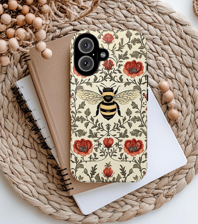 May include: A phone case with a floral and bee design. The case features a cream background with red flowers, green leaves, and a large bee in the center. The phone case is on top of a notebook and a woven mat.