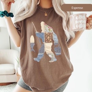 May include: Espresso-colored t-shirt featuring a patchwork Bigfoot design fishing with a fish-shaped lure. The Bigfoot is composed of various fabric patterns, including floral and denim. The shirt has a relaxed fit.