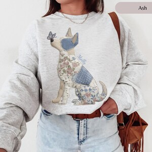 May include: Light grey sweatshirt with a patchwork dog design. The dog has floral and blue patterned patches, with a butterfly on its nose. The name "Molly" is written on the design. The sweatshirt has long sleeves.