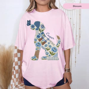 May include: A light pink t-shirt featuring a graphic of a dog silhouette filled with a blue floral and plaid patchwork pattern. A small blue butterfly rests on the dog's nose, and the name Cooper is printed in a cursive script next to the dog.