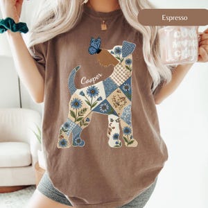 May include: An espresso-coloured t-shirt featuring a large graphic of a dog silhouette filled with a blue and beige floral patchwork pattern. A blue butterfly rests on the dog's nose, and the name Cooper is printed in white script above the tail.