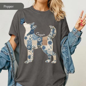 May include: A charcoal grey short-sleeve t-shirt featuring a graphic of a dog silhouette filled with a blue and white floral and plaid patchwork pattern. A blue butterfly rests on the dog's nose, and the name Cooper is written in white script.