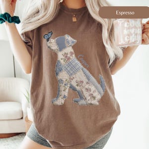 May include: Espresso-colored t-shirt featuring a patchwork dog design with floral and plaid patterns. A blue butterfly rests on the dog's nose. The name "Cooper" is written on the shirt. The shirt is a comfortable, casual style.