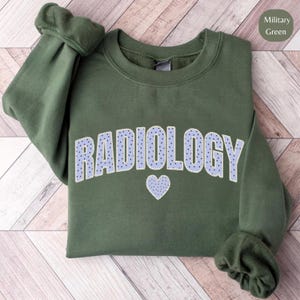 Może przedstawiać: Zielona wojskowa bluza z napisem "RADIOLOGY" w jasnoniebieskich, kropkowanych literach. Małe niebieskie serce znajduje się pod napisem. Bluza ma okrągły dekolt i długie rękawy.