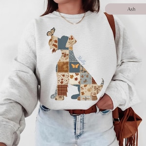 Op de afbeelding: Een asgrijze sweater met ronde hals en een grote hondensilhouet bestaande uit een patchworkpatroon in bruin-, crème- en blauwtinten. Het ontwerp bevat bloem- en bladmotieven, een vlinder op de kop van de hond en de naam Molly in cursief schrift.