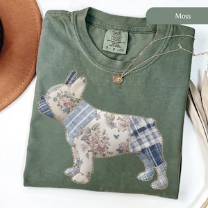 Könnte beinhalten: Ein moosgrünes T-Shirt mit einem Patchwork-Design eines französischen Bulldoggen. Der Hund ist aus floralem und kariertem Stoff gefertigt. Eine goldene Halskette mit Anhänger ist auf dem T-Shirt drapiert. Das T-Shirt hat ein Comfort Colors-Etikett.
