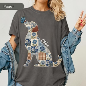 May include: A charcoal gray t-shirt featuring a graphic of a dog silhouette filled with a blue and white floral patchwork pattern. A blue butterfly rests on the dog's nose, and the name Chloe is printed in white script next to the dog's back.