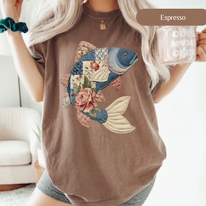 May include: Espresso-colored t-shirt featuring a detailed fish design. The fish is a patchwork of blue, pink, and floral fabrics, with a cream-colored tail. The design includes a blue eye and pink fins. The shirt is a comfortable, casual style.