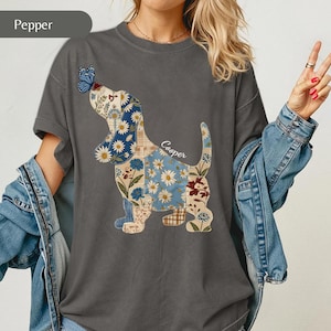 May include: A charcoal grey t-shirt featuring a graphic of a dog in a patchwork style with floral and plaid patterns in shades of blue, white, and brown. A blue butterfly rests on the dog's nose, and the name Cooper is written in white script.
