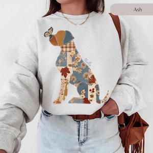 May include: An ash gray crewneck sweatshirt featuring a graphic of a dog composed of various patchwork patterns in shades of blue, brown, and cream, including floral and leaf motifs. A butterfly rests on the dog's nose, and the name Molly is printed.