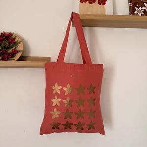 Peut inclure: Un sac fourre-tout corail avec des motifs floraux dorés. Le sac est suspendu à un mur. Le sac fourre-tout a deux longues sangles et une forme rectangulaire. Le motif floral doré est disposé en trois rangées de quatre fleurs.