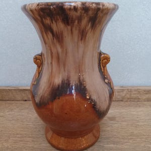 May include: A brown ceramic vase with a wide, flared top and a rounded body. The vase features two small handles and a pedestal base. The color transitions from a dark brown at the top to a lighter brown and orange at the base.
