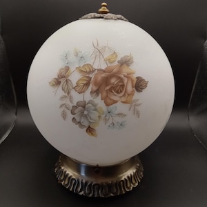 May include: A spherical white glass lamp shade featuring a floral design with brown and light blue roses and leaves. The lamp has a metallic bronze-colored base with a scalloped edge and a small brass-colored finial at the top center.