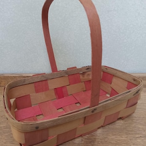 May include: A rectangular basket with a woven design in shades of red and tan. The basket has a curved handle and is made of wood. The basket is sitting on a wooden surface.