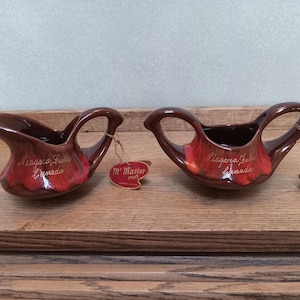 May include: Two brown ceramic gravy boats with a red glaze at the base. Each boat has a handle and is labeled "Niagara Falls Canada" and "M' Master craft". The boats are displayed on a wooden surface.