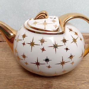 May include: White ceramic teapot with gold accents, including the handle, spout, and lid. The teapot is decorated with gold starburst patterns. A vintage-style tea accessory.