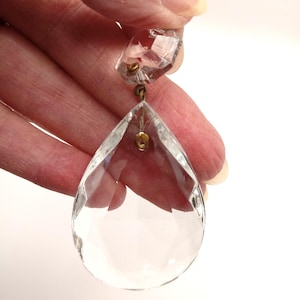 May include: A clear, faceted crystal teardrop pendant with a smaller, geometric crystal above it. The crystals are connected by a small, gold-colored metal loop. The pendant is held against a white background.