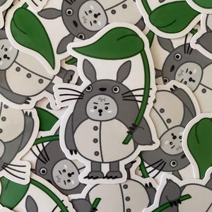 May include: A die-cut sticker featuring a cartoon creature with gray fur, a white belly with three dark gray buttons, pointed ears, and long whiskers. The creature holds a large green leaf over its head like an umbrella, with a surprised expression.