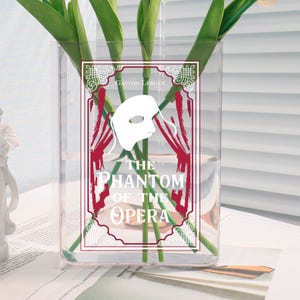 Könnte beinhalten: Eine transparente Glasvase mit einem dekorativen Etikett mit dem Text "The Phantom of the Opera" und einer weißen Maske. Die Vase enthält mehrere weiße Tulpen mit grünen Stielen und Blättern. Eine kleine weiße Engelsstatue und ein Buch sind ebenfalls zu sehen.