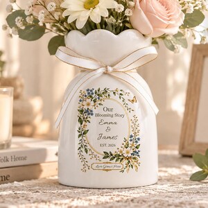 May include: A white ceramic vase with a scalloped rim and a cream-colored ribbon tied around the neck. The front features a floral wreath graphic with the text Our Blooming Story, Emma & James, EST. 2026, and Love Grows Here, filled with white daisies and pink roses.