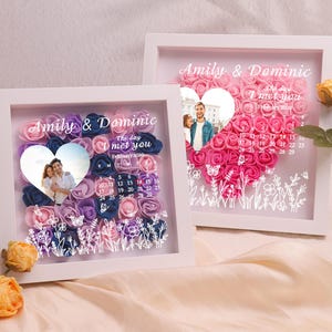 May include: Two white framed shadow boxes with floral heart designs. Each box features a photo, the names "Amily & Dominic", and the phrase "The day I met you". One box has blue, purple, and pink flowers, the other has pink flowers.