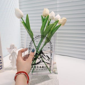 Pu&ograve; includere: Un vaso di vetro trasparente con un design grafico in bianco e nero che presenta montagne, stelle e il testo "Those who listen and the dreams that are answered Velaris City of Starlight". Il vaso &egrave; pieno di tulipani bianchi.