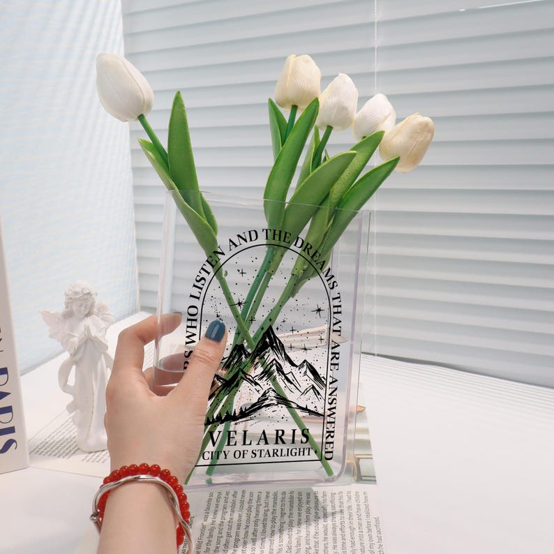 Pu&ograve; includere: Un vaso di vetro trasparente con un design grafico in bianco e nero che presenta montagne, stelle e il testo "Those who listen and the dreams that are answered Velaris City of Starlight". Il vaso &egrave; pieno di tulipani bianchi.