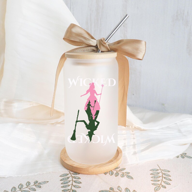 May include: Frosted glass tumbler with a wooden lid, a stainless steel straw, and a tan ribbon. The tumbler features a pink and green silhouette design with the word "WICKED" in white. The tumbler sits on a small wooden coaster.