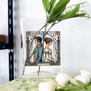 May include: A clear glass vase with a rectangular Pride and Prejudice graphic. The graphic features a man and woman in period clothing, with the text "Pride and Prejudice" and "Jane Austen". The vase holds white flowers.
