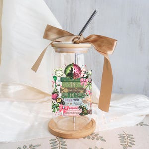 May include: A clear glass tumbler with a bamboo lid and a stainless steel straw. The tumbler features a decorative design with green, pink, and gold accents, including text that reads "Defying Gravity" and "Popular". A gold ribbon is tied around the lid.