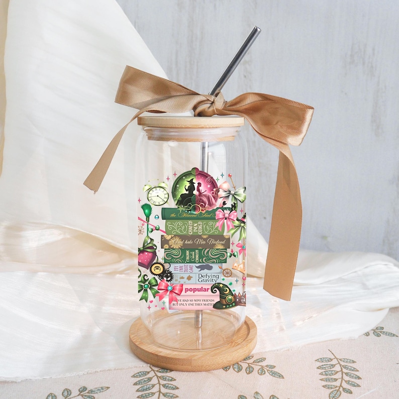 May include: A clear glass tumbler with a bamboo lid and a stainless steel straw. The tumbler features a decorative design with green, pink, and gold accents, including text that reads "Defying Gravity" and "Popular". A gold ribbon is tied around the lid.