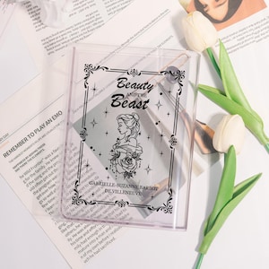Può includere: Custodia in plastica trasparente con un'illustrazione nera di Belle da "La Bella e la Bestia", il testo "Beauty and the Beast" e "GABRIELLE-SUZANNE BARBOT DE VILLENEUVE". La custodia è decorata con motivi floreali.