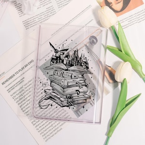 Op de afbeelding: Een transparante, rechthoekige acryl hoes met een zwart-wit illustratie van een stapel boeken, een kasteel en een uil. De hoes is versierd met sterren en een vogel in vlucht. Het woord "Herbology" is zichtbaar op een van de boeken.