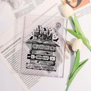 May include: Clear plastic phone case featuring a black and white illustration of a stack of books with the words "Potions", "Herbology", and "Magical Theory". A castle and flying birds are above the books.