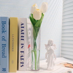 Pu&ograve; includere: Una statuina di angelo in ceramica bianca con ali e una corona, in piedi su un tavolo accanto a un vaso di vetro trasparente pieno di tulipani bianchi e tre libri intitolati "Book of Bread", "La P&acirc;tisserie" e "A Table in Paris".