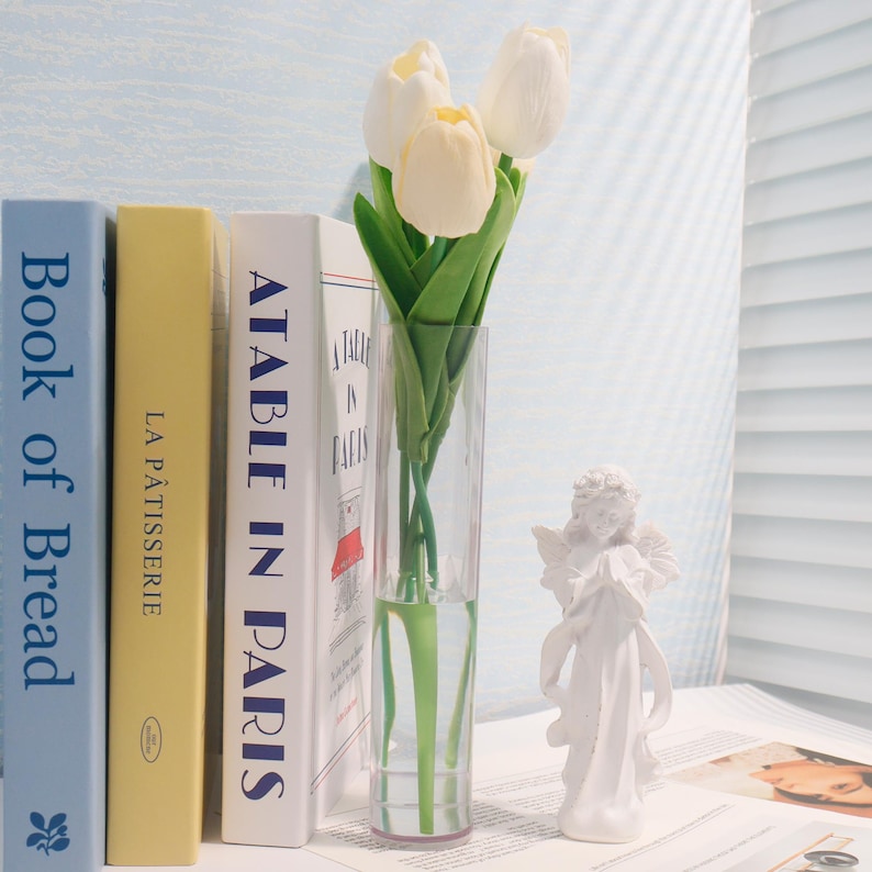 Pu&ograve; includere: Una statuina di angelo in ceramica bianca con ali e una corona, in piedi su un tavolo accanto a un vaso di vetro trasparente pieno di tulipani bianchi e tre libri intitolati "Book of Bread", "La P&acirc;tisserie" e "A Table in Paris".