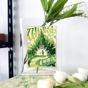 Puede incluir: Un jarrón de cristal transparente con un póster del musical "Wicked" en su interior, con una combinación de colores verde y amarillo que representa la Ciudad Esmeralda. Flores blancas y hojas verdes están dispuestas en el jarrón, con flores blancas adicionales en primer plano.