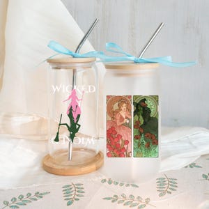 May include: Two glass tumblers with bamboo lids and metal straws. One is clear with a pink and green silhouette and the word "WICKED". The other is frosted with a floral design and images of two women. Both have light blue ribbons.
