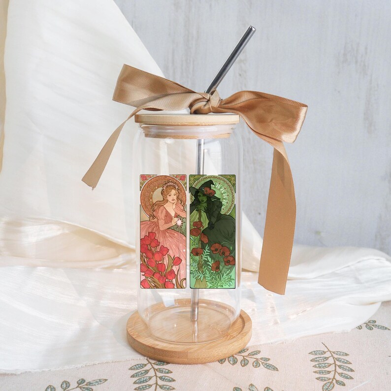 May include: A clear glass tumbler with a wooden lid and base, featuring a stainless steel straw. The tumbler is decorated with a ribbon and a sticker with two Art Nouveau-style illustrations of women in pink and green. The tumbler is designed for cold drinks.