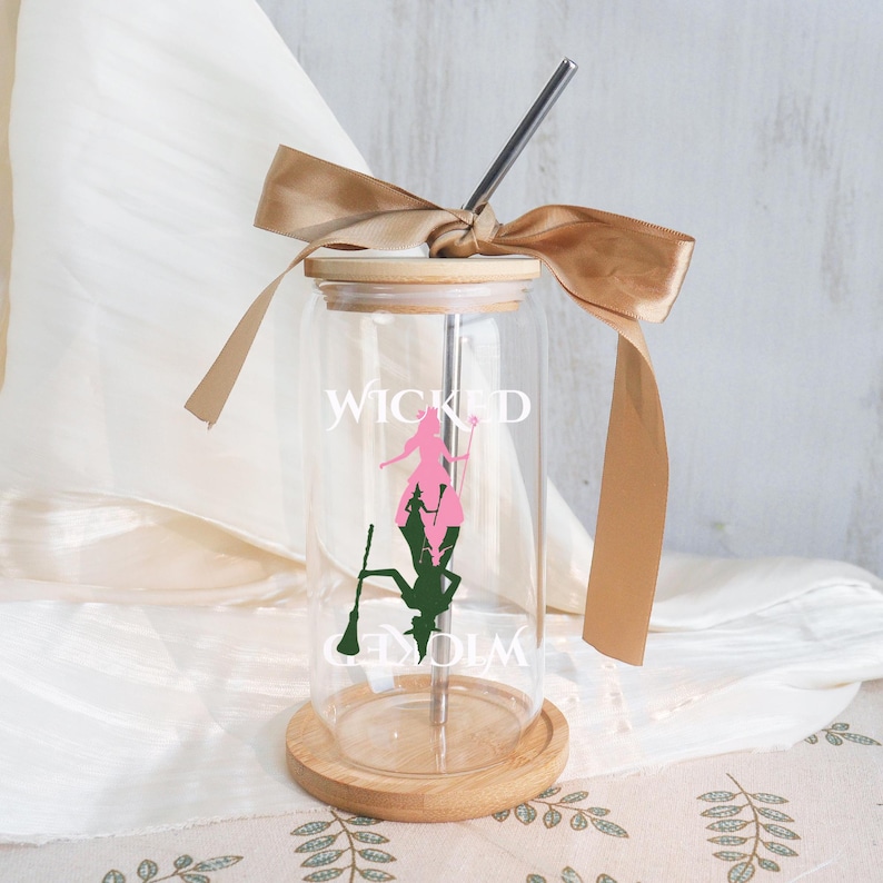 May include: A clear glass tumbler with a wooden lid and base, featuring a stainless steel straw and a tan ribbon tied around the top. The tumbler has the word "WICKED" and a silhouette design in pink and green. The tumbler is designed for cold beverages.