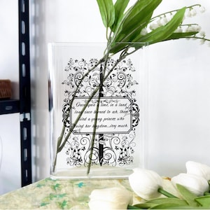 May include: Clear rectangular glass vase with a black and white floral design and text. The text reads "Once upon a time, in a land long since burned to ash, there lived a young princess who loved her kingdom...very much." The vase holds white flowers and green stems.