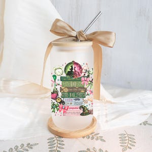 May include: Frosted glass tumbler with a bamboo lid and a stainless steel straw. The tumbler features a colourful design of stacked books, a clock, and a witch's hat. The design includes the words "Defying Gravity" and "Popular". A gold ribbon is tied around the lid.