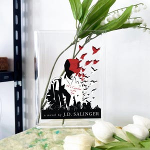Könnte beinhalten: Transparente Glasvase mit einem grafischen Design aus dem Roman "Der Fänger im Roggen" von J.D. Salinger. Das Design zeigt eine Silhouette eines Mannes mit rotem Hut, Vögeln und einer Skyline. Weiße Blumen und grüne Blätter sind sichtbar.