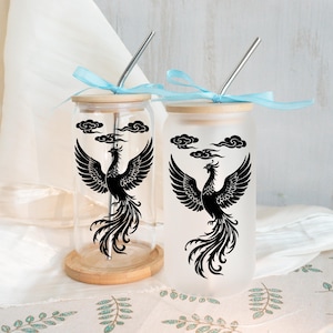 Puede incluir: Dos vasos de vidrio con tapas de bambú y pajitas de acero inoxidable. Cada vaso presenta un diseño de fénix negro con alas extendidas y un motivo de nube. Los vasos están atados con cintas azul claro.